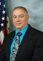 Photo of David Truax wearing a necktie, button shirt and a black blazer with the American flag and a light blue background