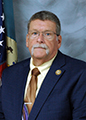 Photo of Jerry Brennan wearing a navy blaze, with a pin on the lapel with the American flag in and a light blue background